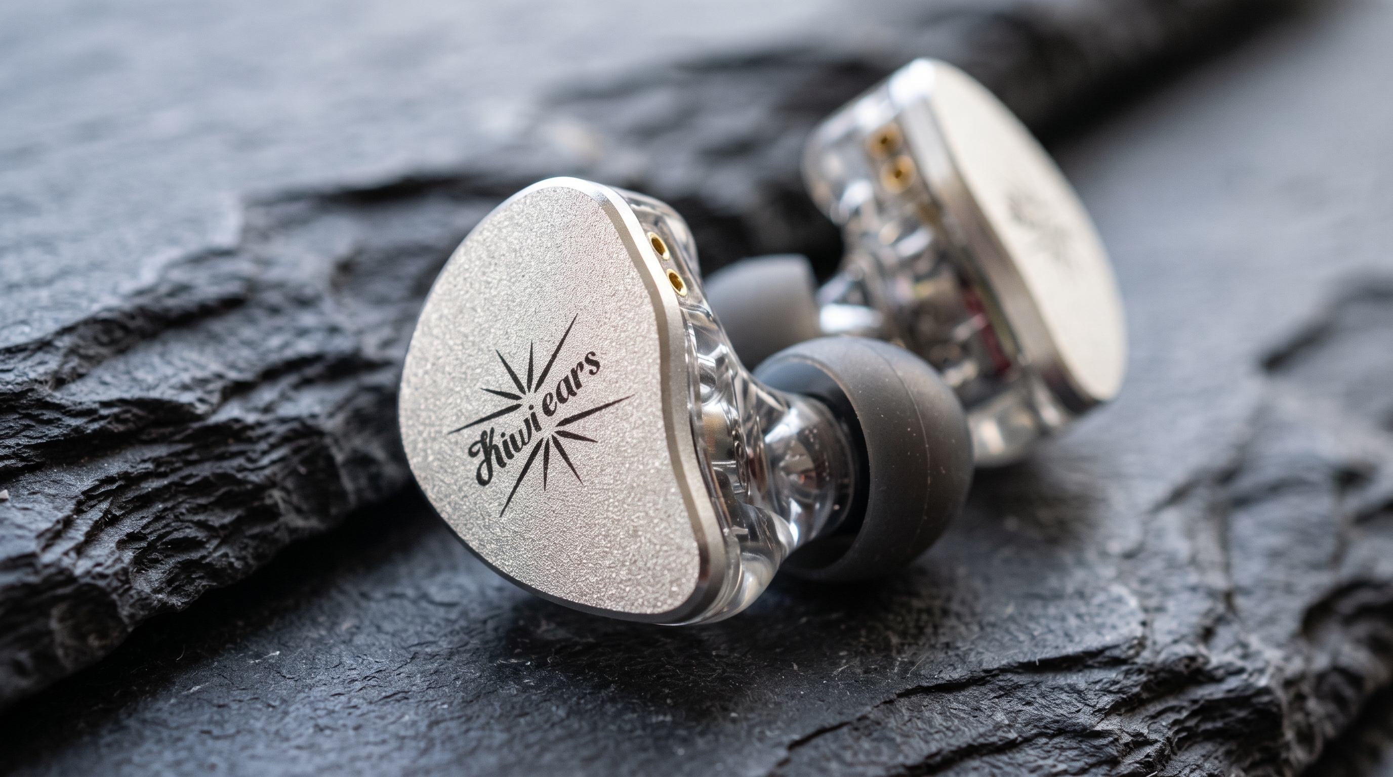 Primer plano macro de las carcasas de los auriculares Kiwi Ears Belle sobre roca pizarra mostrando la textura de la placa metálica y la transparencia de la resina